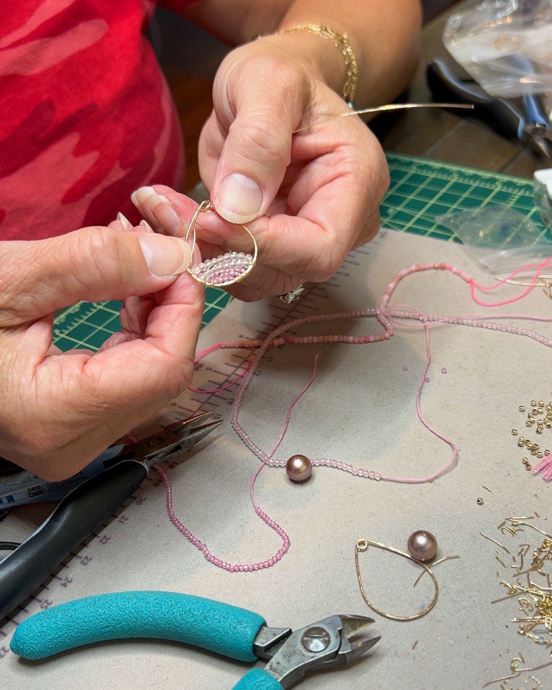 Close-up of hands crafting a pair of gold and pink beaded earrings, surrounded by jewelry tools and materials on a workbench.
