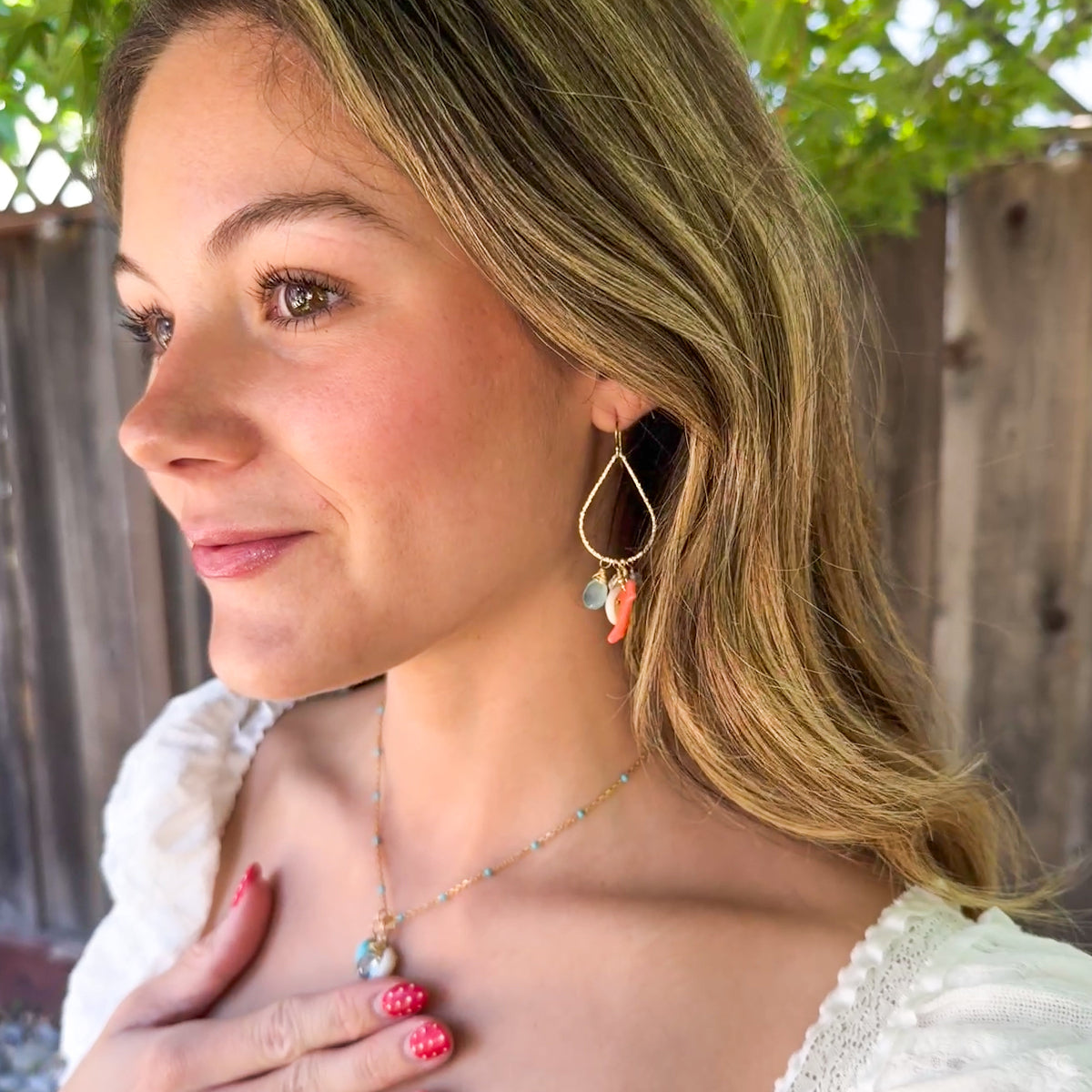 Woman wearing teardrop hoop earrings with puka shells, coral & chalcedony gemstones